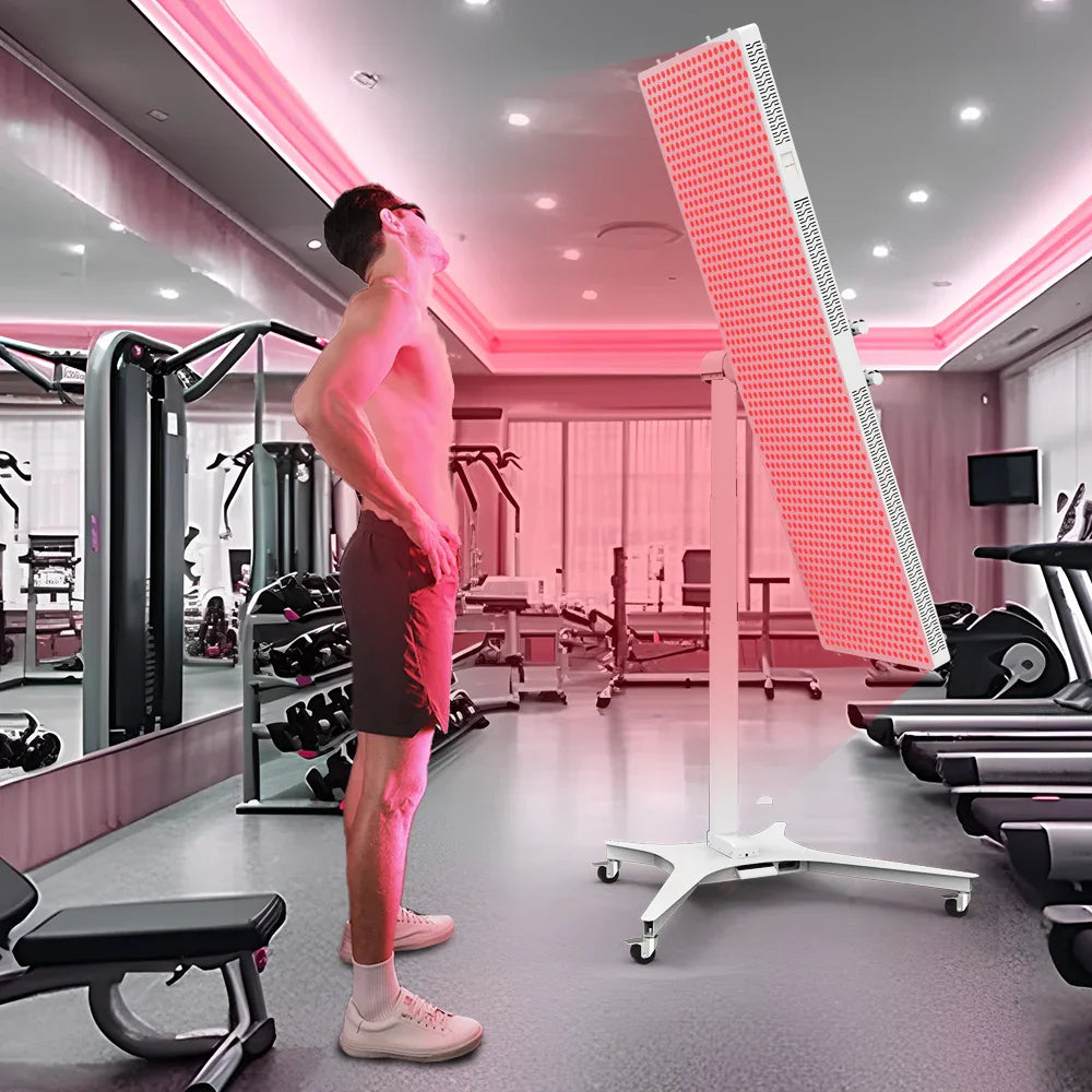 Man standing shirtless in gym under red light therapy panel with exercise machines