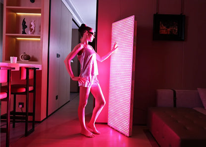 Woman in pajamas using large red light therapy panel in modern living room