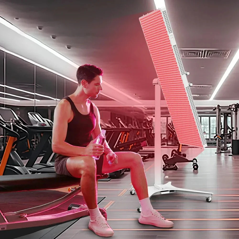 Man sitting on gym bench holding water bottle near red light therapy panel in modern fitness center