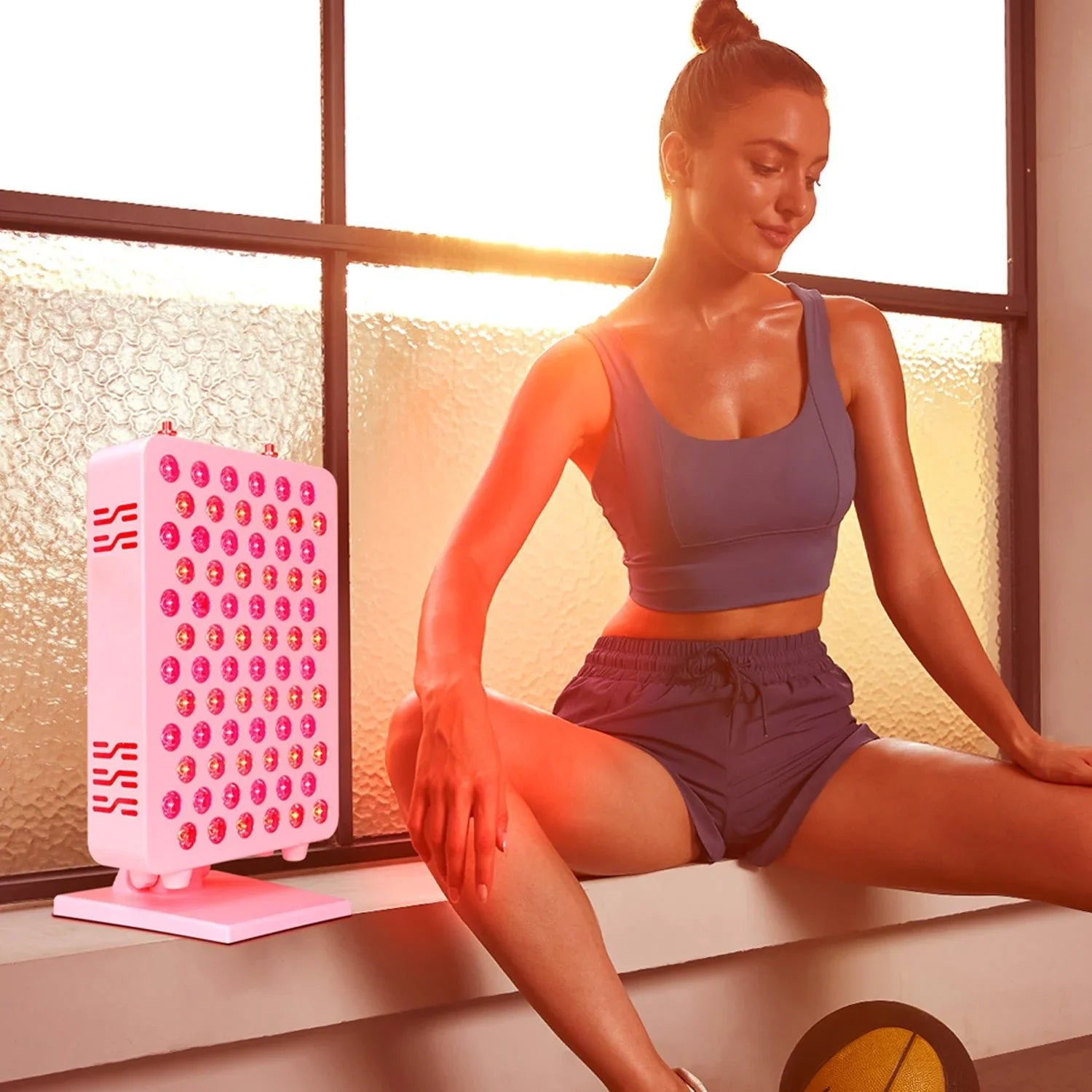 Woman in workout outfit using red light therapy panel indoors by frosted window