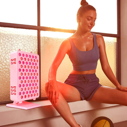 Woman in workout outfit using red light therapy panel indoors by frosted window